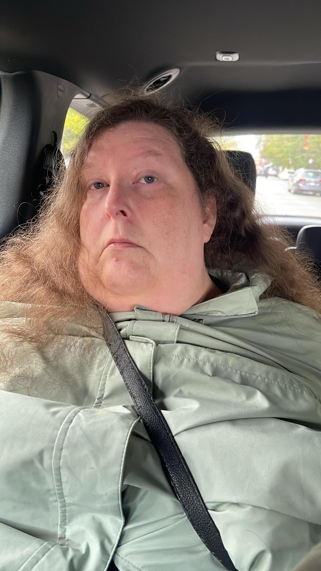 Woman with long, wavy hair wearing a light green jacket sits in a car, looking pensively out the window.