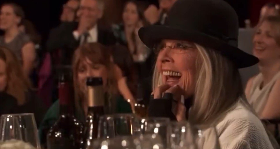 Diane Keaton smiling and laughing at an event, surrounded by an audience and wine bottles, showcasing a joyful atmosphere.