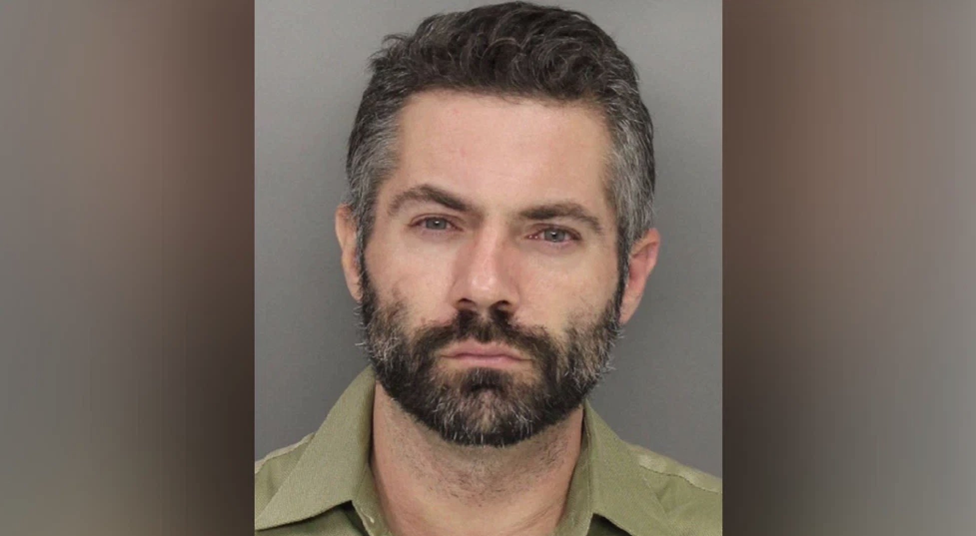 Mugshot of a man with a beard and short hair, wearing a green shirt, looking directly at the camera against a gray background.